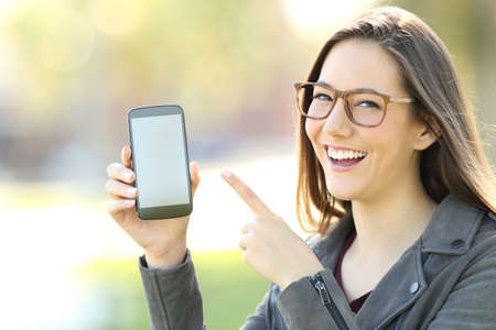 Happy woman wearing eyeglasses showing a blank phone screen in the streetの写真素材