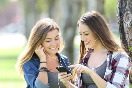 Two happy teen friends listening music sharing earphones and smartphone in a parkの写真素材