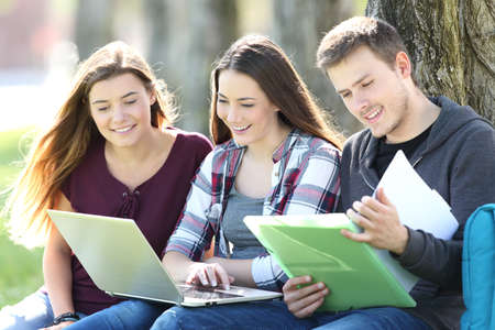 Three happy students studying on line with a laptop and reading notes sitting on the grass in a park