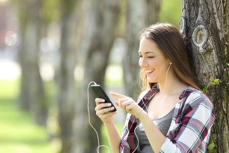 Beautiful teen girl listening and choosing music in a smart phone leaning on a tree in a parkの写真素材