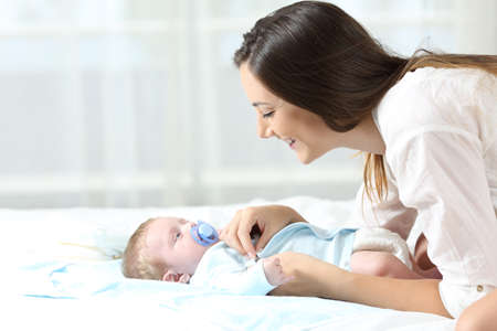 Side view portrait of a happy mother dressing her baby son on a bed at homeの写真素材