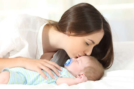 Side view portrait of an affectionate mother kissing her baby sleeping on a bedの写真素材