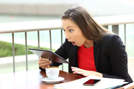 Amazed executive receiving good news in a tablet sitting in a coffee shopの写真素材