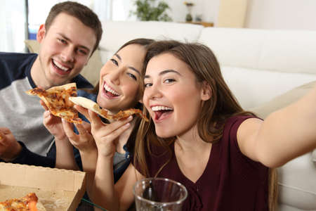 Three happy friends taking selfies and eating pizza sitting on a sofa at homeの写真素材