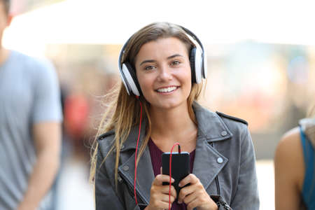 Front view portrait of a happy girl listening to music on phone and looking at you on the streetの写真素材