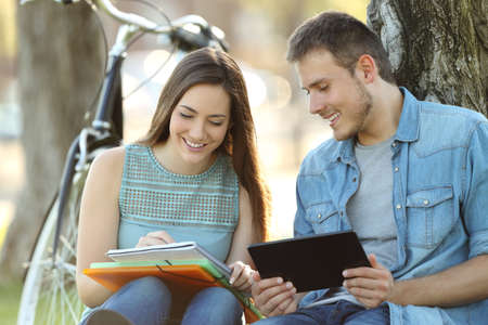 Two happy students learning together on line with a tablet and taking notes sitting in a parkの写真素材