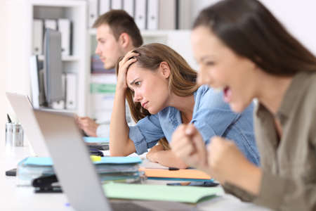 Frustrated employee beside successful one reading news on line sitting in a desktop at officeの写真素材
