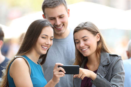 Three happy friends sharing media content with a smart phone on the streetの写真素材