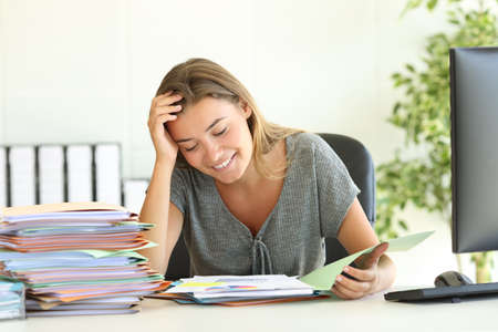 Front view portrait of a happy employee reading report sitting in a desktop at officeの写真素材
