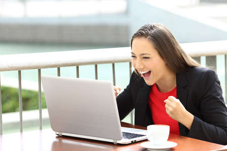 Excited executive reading an on line notification in a laptop sitting in a coffee shopの写真素材