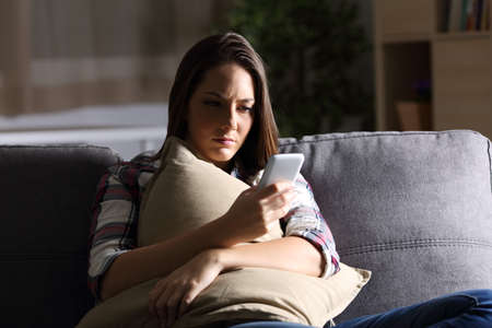 Sad girl holding pillow and reading phone message in the dark sitting on a couch at homeの写真素材