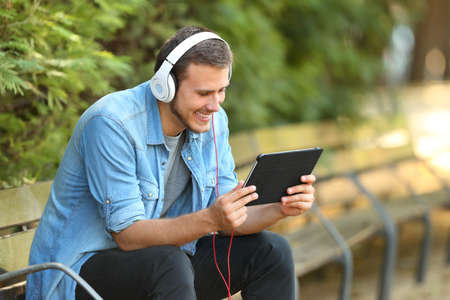 Portrait of a happy guy relaxing watching videos on line in a tablet sitting on a bench in a parkの写真素材