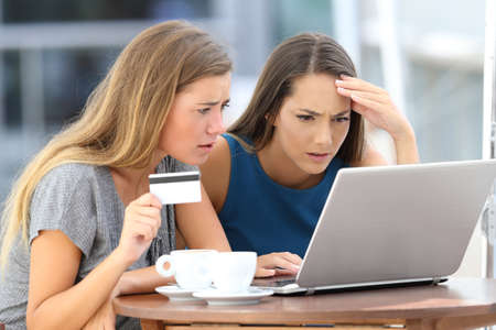 Two worried women having problems buying on line with a laptop and credit card sitting in a barの写真素材