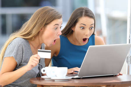 Two astonished friends buying on line with a laptop and credit card sitting in a bar terraceの写真素材
