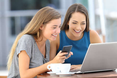 Two happy friends using a laptop and a smart phone sitting in a bar terraceの写真素材