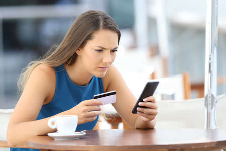 Single worried girl having problems paying on line with a credit card and a phone in a coffee shop terrace