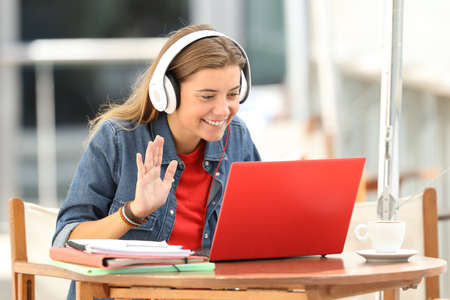 Happy student having a video conference with a red laptop and headphones sitting in a bar terraceの写真素材