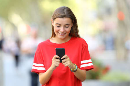 Front view of a happy teen using a smart phone walking on the streetの写真素材