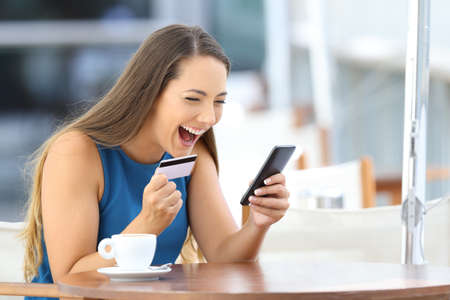 Excited on line buyer paying with credit card using a smart phone sitting in a bar terraceの写真素材