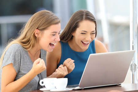 Two excited friends finding on line content in a laptop sitting in a restaurant terrace