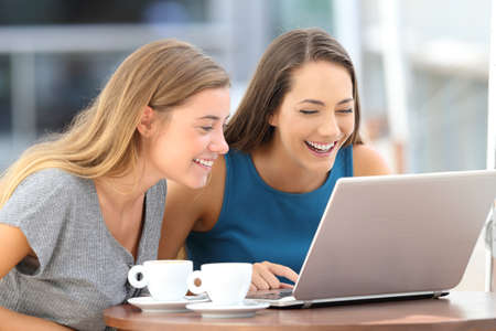 Two happy friends laughing watching on line content in a laptop sitting in a restaurant terraceの写真素材