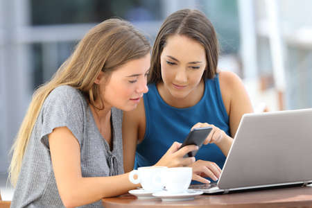 Two friends sharing on line information with a phone and a laptop sitting in a restaurant terraceの写真素材