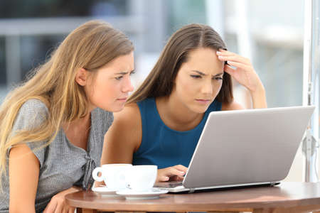 Two worried friends watching media content on line in a laptop sitting in a restaurant terraceの写真素材