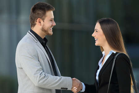 Side view of two happy executives meeting and handshaking on the street with an office building in the background