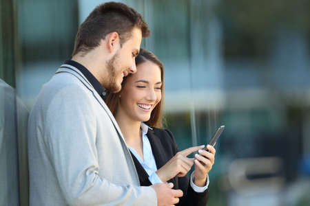 Two executives using a mobile phone leaning on a business building on the streetの写真素材