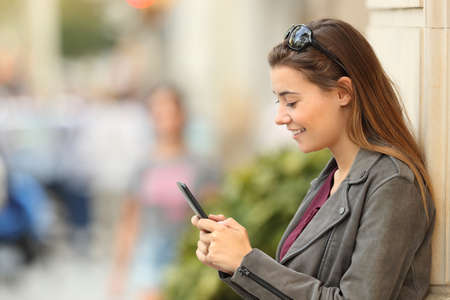 Profile of a fashion girl using a smart phone standing outside leaning on a wall on the streetの写真素材