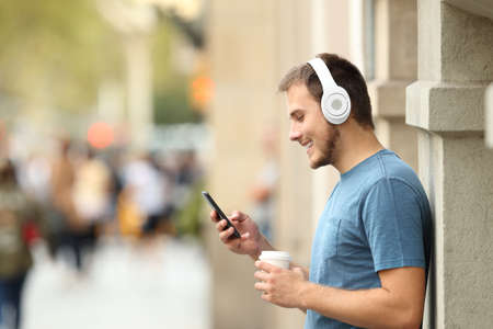 Profile of a happy guy listening music on line with headphones and a smart phone leaning on a wall on the streetの写真素材