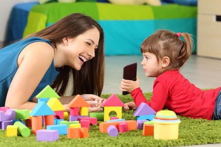 Toddler showing a smart phone to her mother lying on a carpet in a roomの写真素材