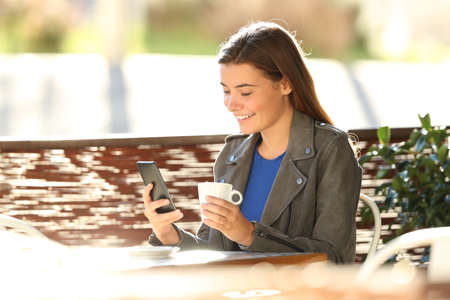 Single fashion teen using a smart phone sitting in a coffee shop terrace with a back light in the backgroundの写真素材