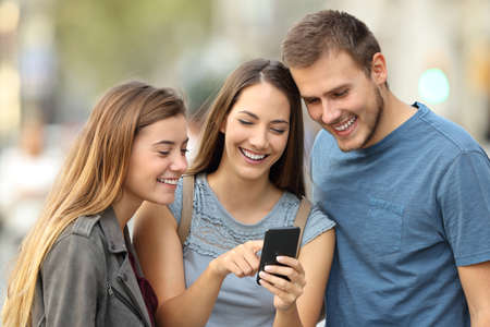 Happy group of three friends using a smart phone standing outdoors on the streetの写真素材