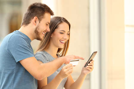 Happy couple buying on line with a smart phone and credit card in front of a storefront on the streetの写真素材