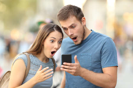 Amazed couple watching on line media in a smart phone standing on the streetの写真素材