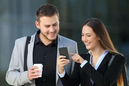 Front view of two happy executives talking about smart phone content standing outdoors on the streetの写真素材