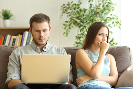 Husband using a laptop on line ignoring to his sad wife sitting on a sofa at homeの写真素材