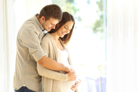Proud new parents looking at belly of pregnant mother standing beside a window at homeの写真素材