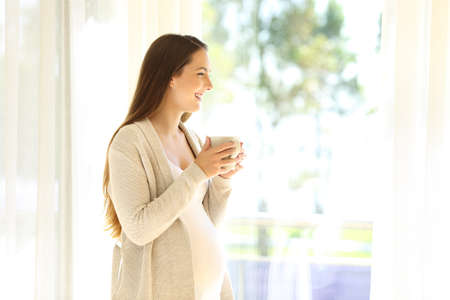 Portrait of a happy pregnant mother looking away through a window at home holding a hot drinkの写真素材