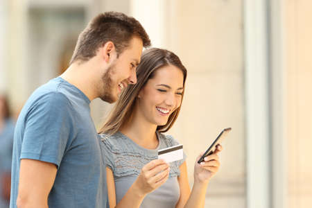 Shoppers buying on line with credit card and smart phone standing beside a storefront on the street