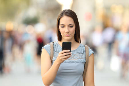 Front view of a serious girl checking a phone message walking on the streetの写真素材