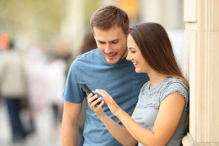 Happy casual couple checking content in a smart phone standing on the streetの写真素材