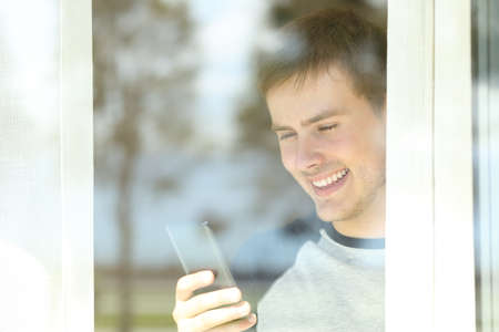 View through a window of a happy man using a smart phone at homeの写真素材