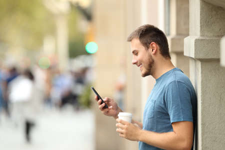 Side view of a happy man using a smart phone and holding a take away drink on the streetの写真素材