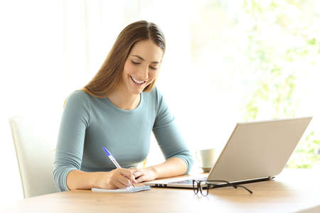 Happy woman writing notes beside a laptop on a desk at homeの写真素材