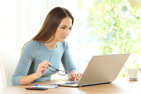 Serious woman holding eyeglasses reading on line content in a laptop on a table at homeの写真素材
