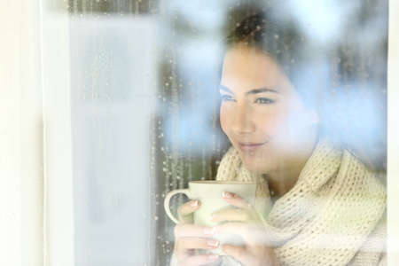 Portrait of a girl looking through a window holding a coffee cup in a rainy day of winterの写真素材