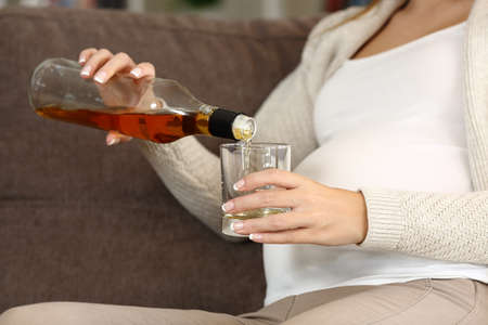 Close up of an irresponsible pregnant woman drinking alcohol sitting on a couch at homeの写真素材