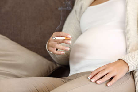 Close up of an irresponsible pregnant woman smoking a cigar sitting on a sofa at homeの写真素材